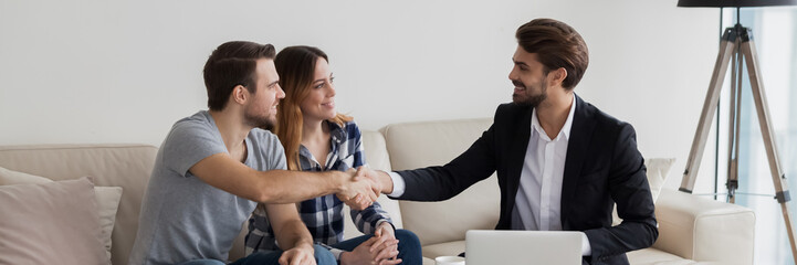 Happy couple young family flat renters shake hands with realtor seated on sofa in living room. Make...