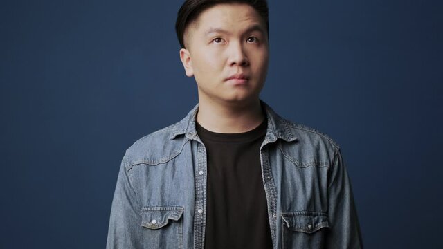 A Confused Young Asian Man Wearing Casual Clothes Is Raising His Shoulders Standing Isolated Over Dark Blue Wall Background In Studio