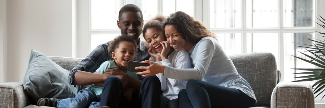 Full African family with little kids sit on couch in living room having fun using smart phone new cool application, taking selfie, watch funny videos. Horizontal photo banner for website header design