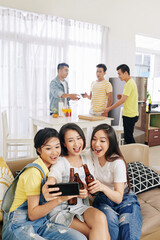 Pretty young Vietnamese women clinking beer bottles and talking selfie at house party