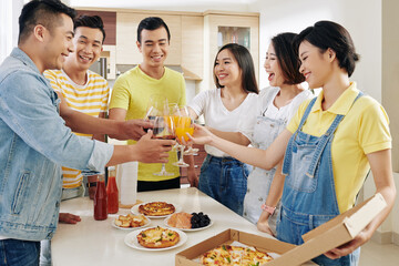 Cheerful friends toasting with glasses of juice over table with tasty pizza and snacks