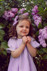 Fototapeta premium three-year-old girl stands in lilac bushes in a dress and a bow