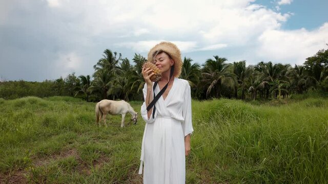 Girl with a pineapple in hand, in a white dress and a straw hat stands in a field