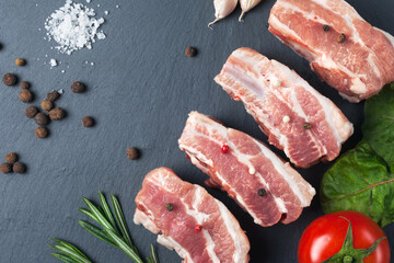 Raw slices of pork on a black slate board, tomatoes, rosemary, garlic, salt and powder. Pork belly with vegetables.