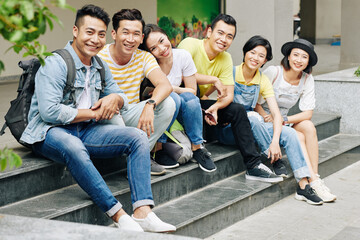 Joyful young Asian people sitting on steps of college or university building and looking at camera © DragonImages