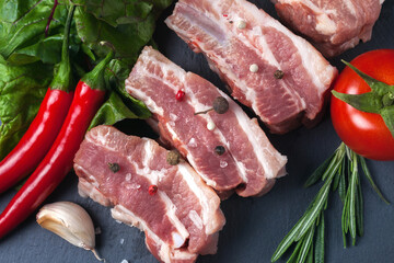 Raw slices of pork on a black slate board, green salad, tomatoes, rosemary, garlic, red pepper and powder. Pork belly with vegetables.