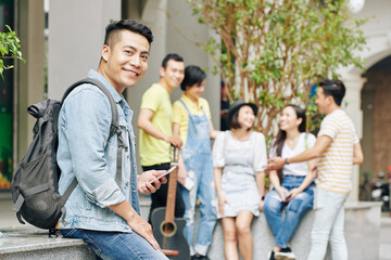 Portrait of handsome young Vietnamese university student with backpack texting and lookng at camera