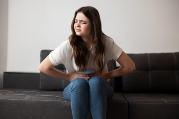 Obraz premium young beautiful brunette woman sitting on the sofa suffering from extreme pain in stomach looking desperate, healthcare and medical concept