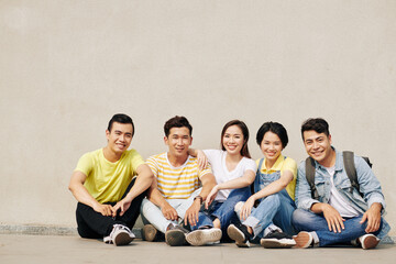 Group of cheerful students sitting on ground next to and smiling at camera