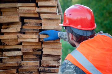 Builder in a protective helmet holds wooden planks. Concept - sale of lumber. Builder is considering wood planks. Concept - wood processing business. wood timber stack of wooden planks.