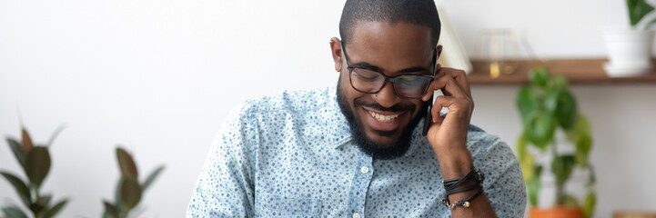 African millennial man holding smartphone enjoy distant conversation seated indoors, horizontal...