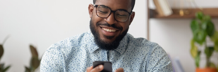 Smiling African businessman sit at workplace holding mobile phone, texting sms, browsing internet....