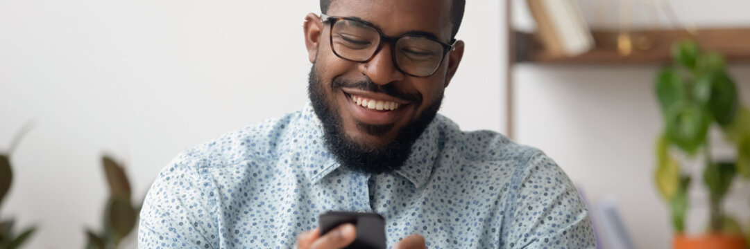 Smiling African businessman sit at workplace holding mobile phone, texting sms, browsing internet. Distant communication, virtual chat usage concept. Horizontal photo banner for website header design