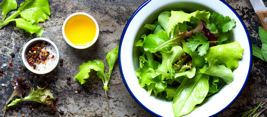 Various lettuce in a bowl, olive oil, salt and pepper on a dark background, top view, banner. Cooking salad dressing with oil and spices. Diet, healthy or vegetarian food.