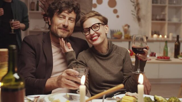 Romantic couple holding glasses with wine, embracing, chatting and then looking at camera and smiling at home dinner party with friends