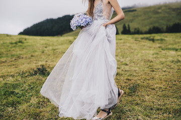 Fototapeta na wymiar a young beautiful girl in a white dress looking like a bride walking through green meadows and hills with gartensia in her hands