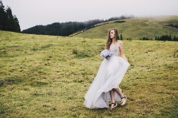 a young beautiful girl in a white dress looking like a bride walking through green meadows and hills with gartensia in her hands