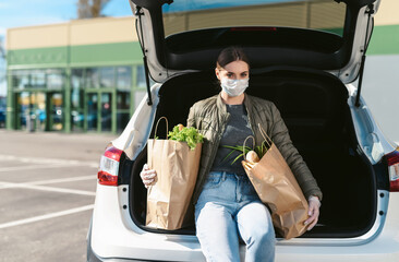 A young woman sitting in a car trunk with groceries from a supermarket. Social distancing: face mask, disposable gloves to prevent infection. Food shopping during coronavirus Covid-19 quarantine