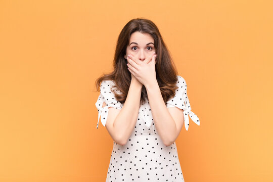 young pretty woman covering mouth with hands with a shocked, surprised expression, keeping a secret or saying oops against orange wall