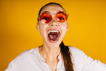 Cheers Cheers !! Girl screams with her mouth wide open and looking through pink glasses on a yellow...