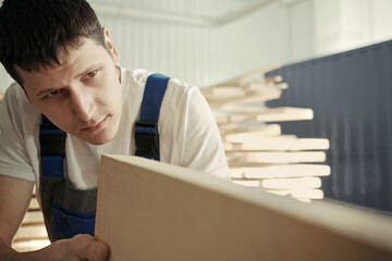 A young carpenter chooses a wooden board. Inspection and quality control of wood. Worker in the carpentry shop.