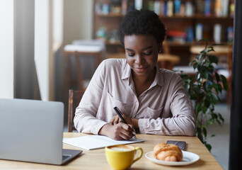 Fototapeta na wymiar Studying online. Young African American girl with laptop taking notes while having breakfast in cafe