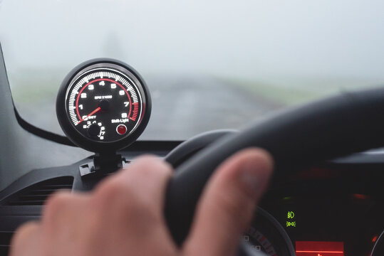 Driver hand on the steering wheel and tachometer with shift light close up on foggy road background.