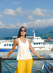 Fototapeta na wymiar Positive woman walking at the harbor on vacation. Mountains on background. Cheerful, smiling, lucky woman in white blouse, yellow trousers and sunglasses, outdoors at sea port. Sunny summer day.