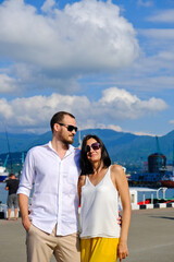 Happy couple of lovers walking at the harbour. Mountains at background. Cheerful, smiling man in white and woman in white blouse and yellow trousers, outdoors at sea port.