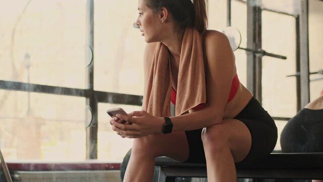 Young concentrated optimistic fitness woman in gym using mobile phone looking at watch clock