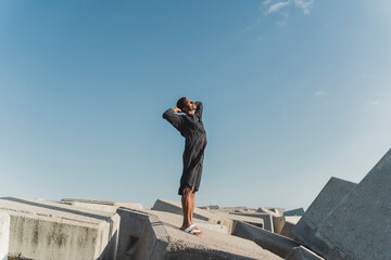 Young man wearing black kaftan standing on concrete blocks under blue sky