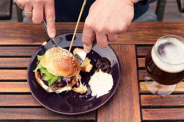 Burger on a plate and hands with knife cutting the hamburger. Healthy fast food in restaurant with vegetables and beer.