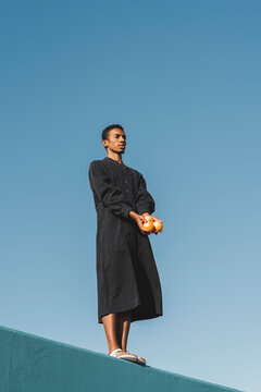 Young man wearing black kaftan standing on blue wall holding oranges