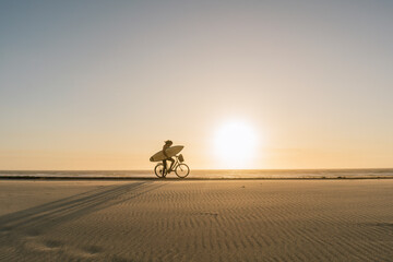 Surfer riding a bicycle during the sunset in the beach, Costa Nova, Portugal