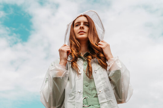 Portrait of redheaded young woman wearing transparent rain coat standing against cloudy sky
