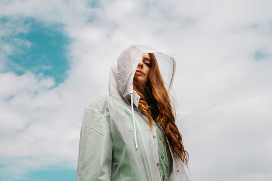 Redheaded young womanwearing transparent rain coat standing against cloudy sky