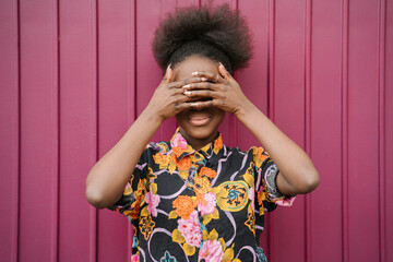 Smiling young woman weaning blouse with floral design covering eyes with her hands