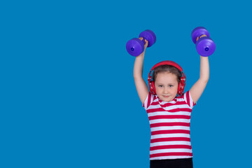 A beautiful little girl with headphones is doing sports with dumbbells in her hands. Model on a blue background