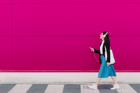 Young woman walking along pink wall, using smartphone and listening to music