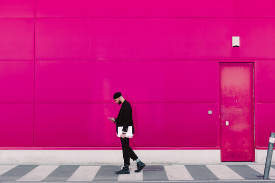 Businessman with smartphone and documents walking along a pink wall