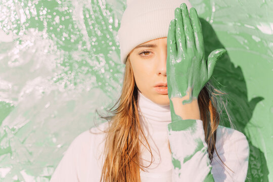 Young woman standing in front of painting, covering eye with green hand