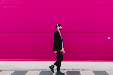 Young man with headphones and smartphone walking along a pink wall