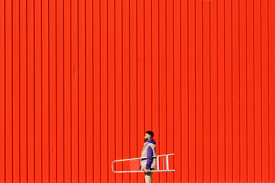 Young Man Carrying A Ladder In Front Of A Red Wall