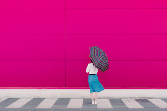 Rear view of young woman with umbrella standing in front of a pink wall