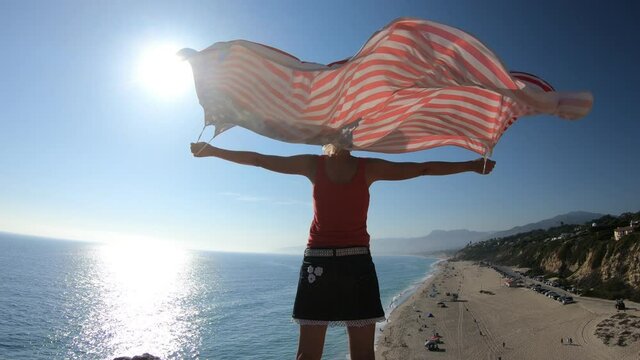 Woman holding an American flag looking Point Dume State Beach from Point Dume promontory on Malibu coast in CA, United States. Carefree woman in California West Coast. Blue sky, sunny.