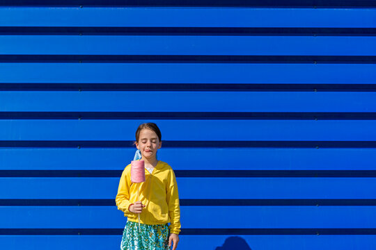 Little girl with fake ice cream standing in front of blue background licking lips
