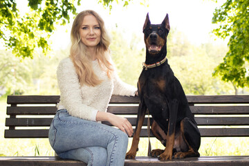 A woman and her dog Doberman sit together on a bench in the park, a girl with a Doberman