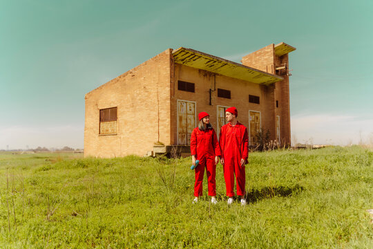 Young couple wearing red overalls and hats standing on a meadow in front of industrial building