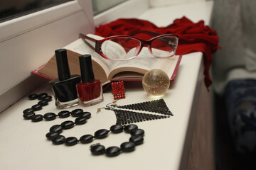 Composition of red glasses, black beads made of natural agate, nail polishes, shiny disco earrings, a transparent shiny ball, an open dictionary, a matrix ring, on a white windowsill.