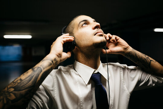 Young businessman with tattoo on his forearms with headphones in a gloomy car park